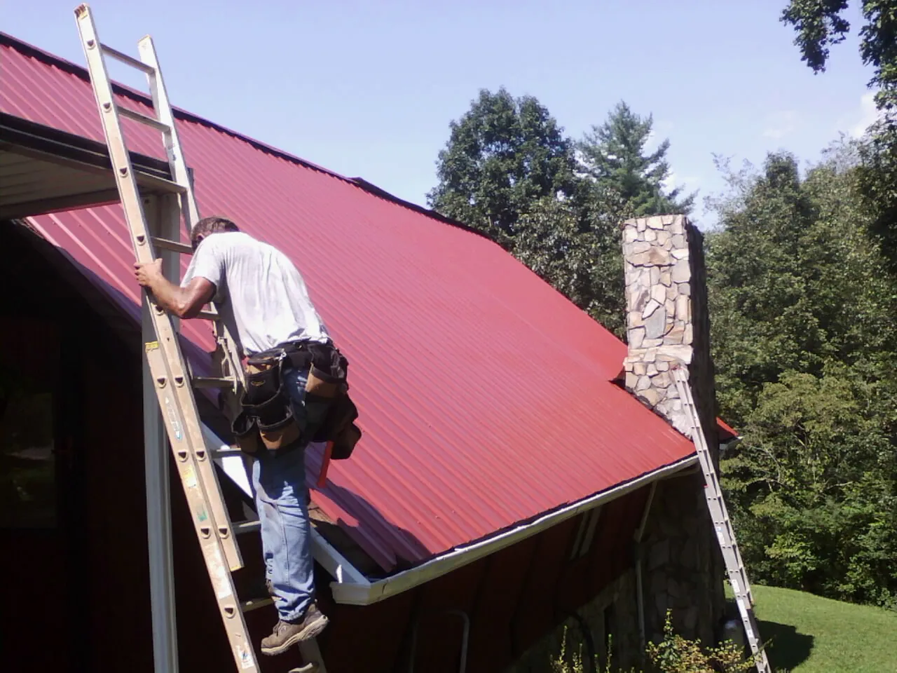 Roofer on a ladder beside a red metal roof and stone chimney — Soffit & Fascia Repair by Roofing Brothers in Morganville